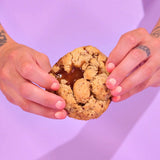 Salted Caramel Pecan New York Cookie with gooey salted caramel centre and toasted pecans held by hands against purple background